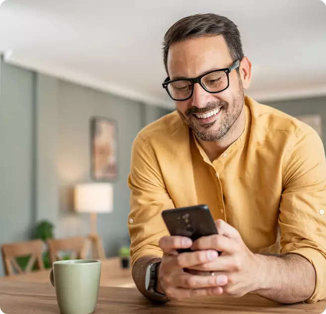 Ein Mann mit Brille und gelbem Hemd lächelt, während er auf sein Smartphone schaut. Im Vordergrund steht eine grüne Tasse auf einem Tisch.