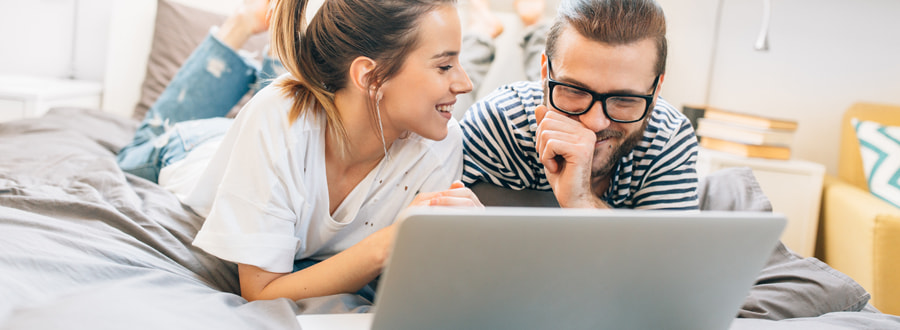 Budgetrechner, junges Paar liegt mit dem Laptop auf ihrem Bett und lächelt, Foto: Astarot/stock.adobe.com